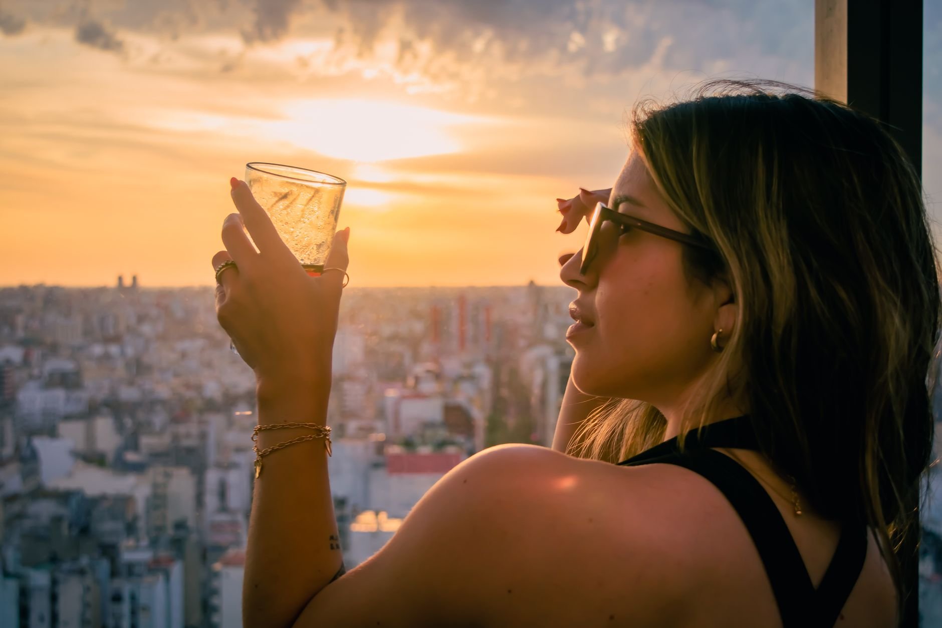 Woman holding a glass, admiring a sunset from a rooftop, cityscape view.